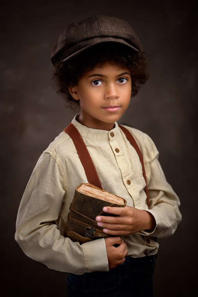 fine art portrait of a african boy wearing a hat and holding a book in vintage setting in san diego studio